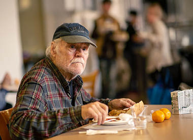 Elderly man eating a meal at a table, with other people in the background